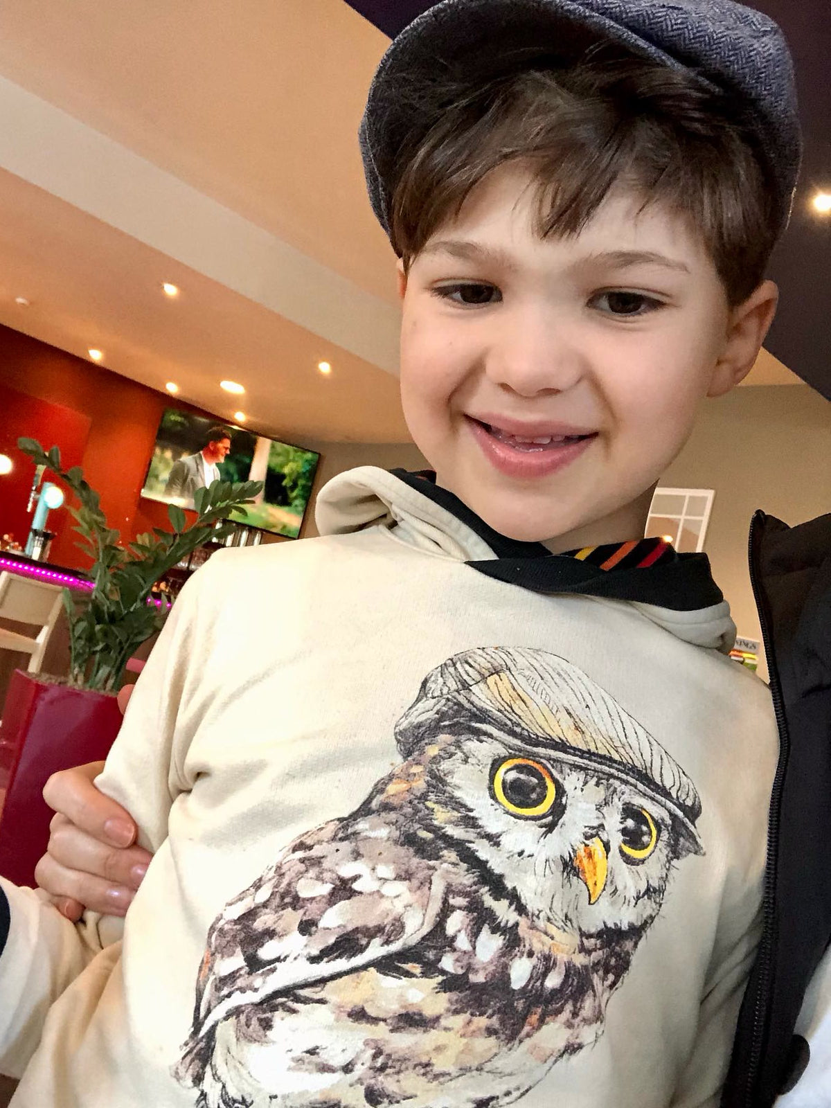 Smiling boy in a flat cap wearing The QT Hatted Hootie organic cotton hoodie, matching the owl in its cap